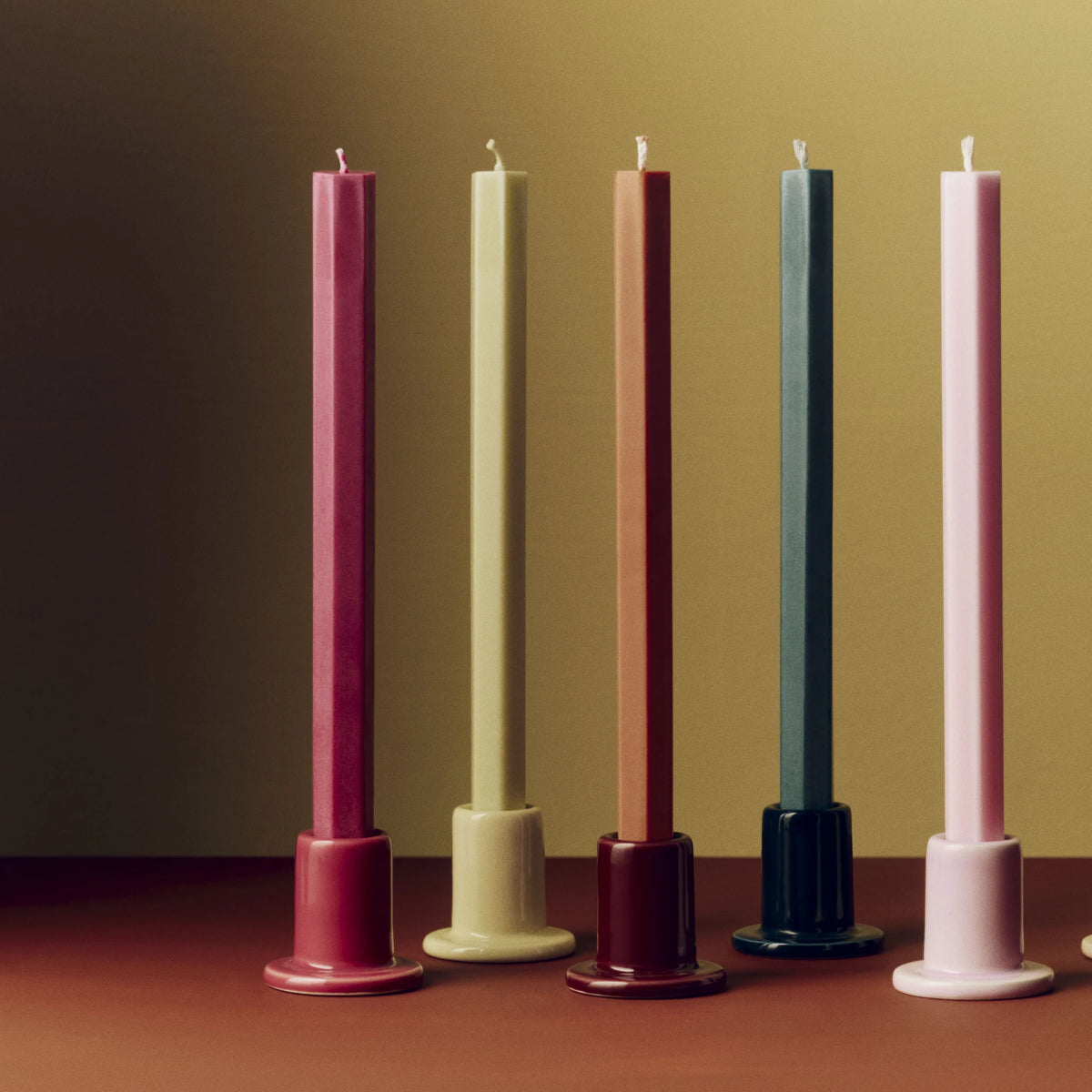 Een rij Hay Tube Kandelaars met lange kaarsen op een terracotta tafel tegen een okergele muur, met de Hay Tube Kandelaar Limoenwit tussen bessenroze en kastanjebruin.