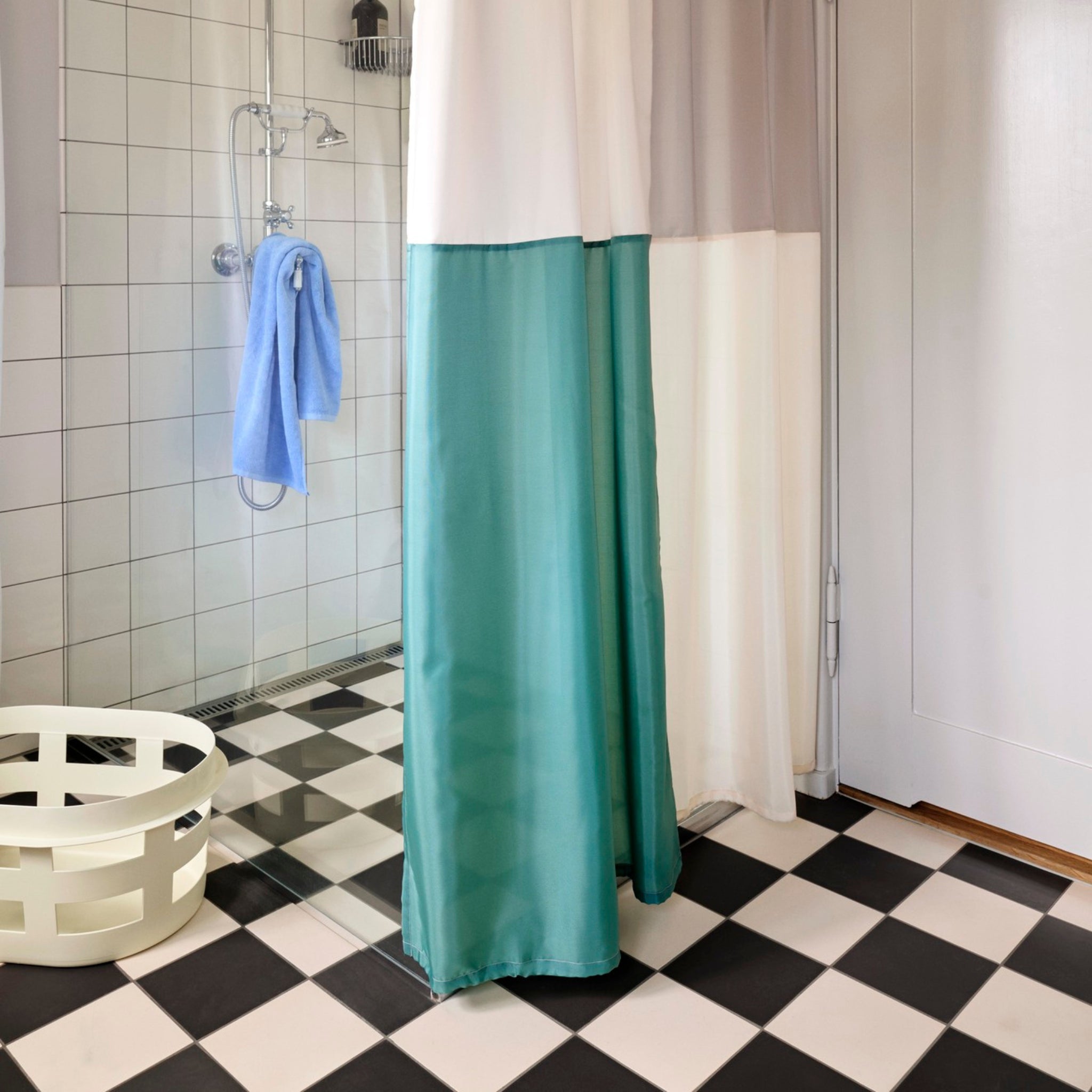Een badkamer met een zwart-wit geblokte vloer, wit betegelde douchewanden, een blauwe handdoek, een wit en groen douchegordijn en een Hay Wasmand Geel wasmand van Hay.