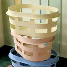 Drie moderne, rechthoekige Hay Wasmand Geel wasmanden van Hay - pastelgeel, roze en blauw - staan netjes gestapeld tegen een lichtgroene muur en een donkerkleurige vloer.