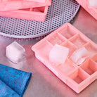 Een Hay ijsblokjesvorm Vierkant XL Lichtblauw zit op een metalen oppervlak bij een blauw gedessineerd doek, met extra lichtblauwe bakjes en een geperforeerd rond dienblad op de achtergrond.