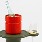 Een heldere wijnfles staat in een gedurfde Heller Asti Ijsemmer Oranje, naast een halfvol glas op groen, met een witte achtergrond-een eerbetoon aan de iconische ontwerpstijl van Sergio Asti.