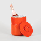 Een fel oranje Heller Asti Ijsemmer Oranje ijsemmer van Heller, geïnspireerd door Sergio Asti, biedt plaats aan ijs en een fles rosé wijn. Het deksel leunt tegen zijn zijkant op een eenvoudige lichtgrijze achtergrond.