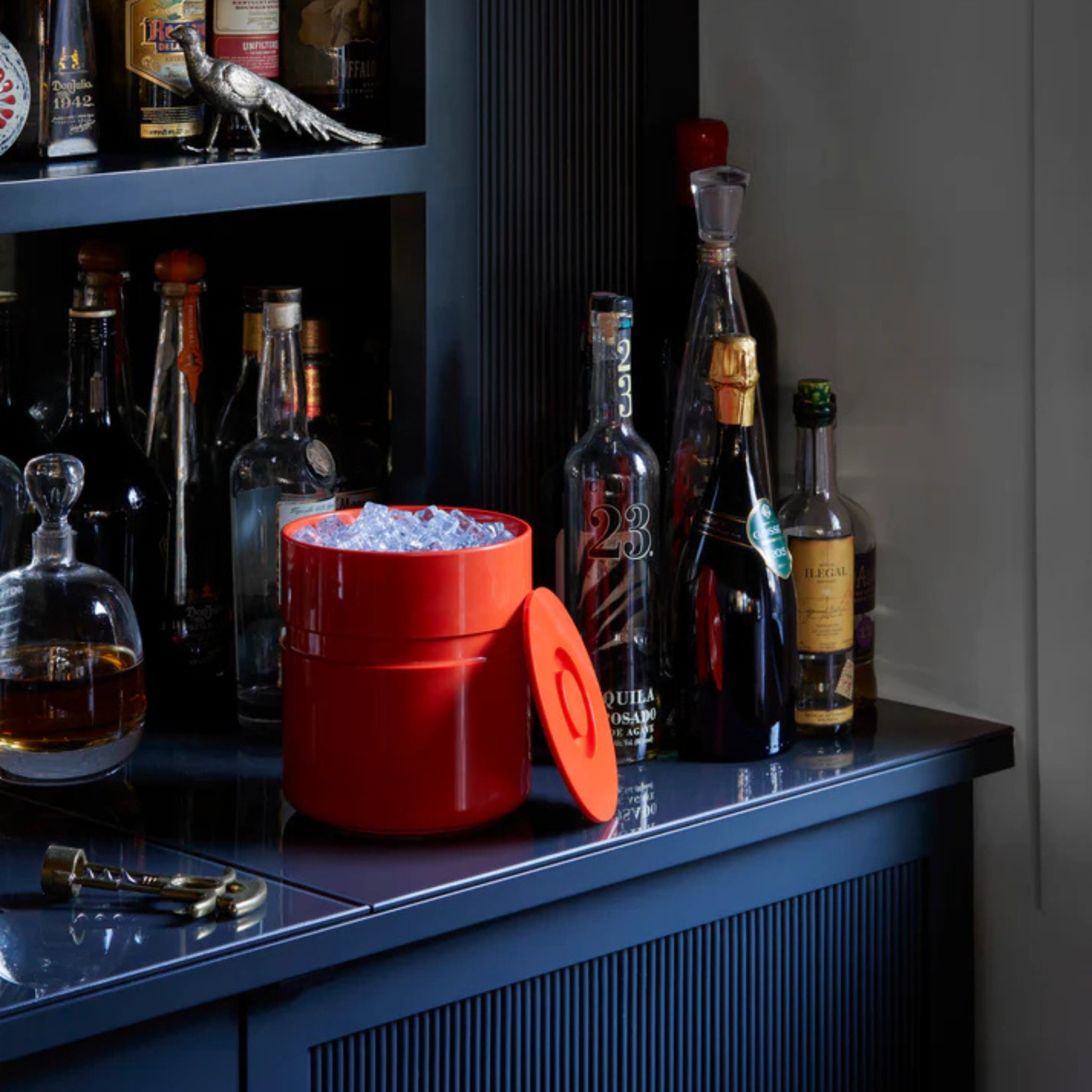 Een Heller Asti Ijsemmer Oranje gevuld met ijs staat op een donkerblauwe bar naast likeurflessen, een kurkentrekker en een karaf, met het deksel vlakbij. Op een plank op de achtergrond staan nog meer flessen, die de tijdloze stijl van Heller oproepen.