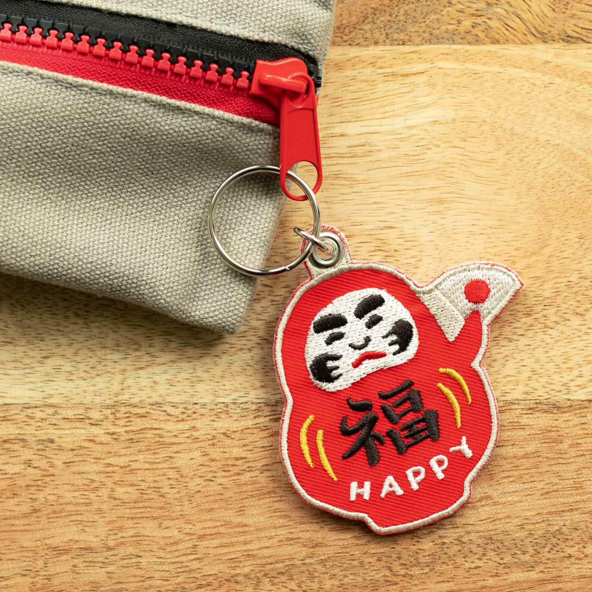 De Hightide Stitch Work Sleutelhanger Daruma, een rode sleutelhanger met "HAPPY" en een kanji-teken, is bevestigd aan een beige etui met een rode rits op hout. Dit Hightide accessoire brengt charme en geluk aan je spullen.