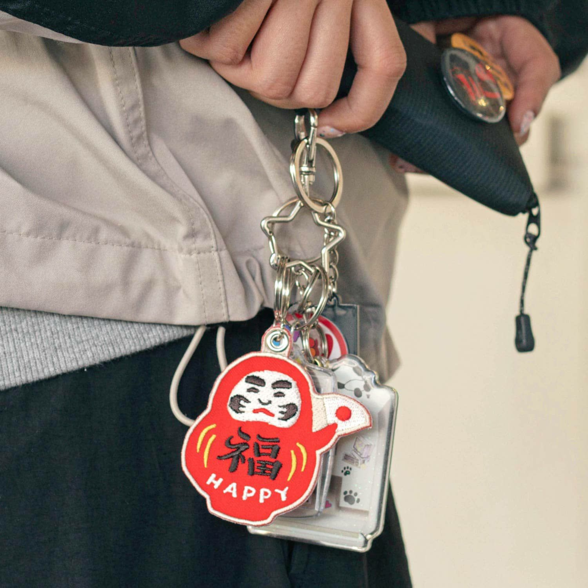 Een persoon houdt een etui vast met de Hightide Stitch Work Sleutelhanger Daruma van Hightide, een rode sleutelhanger die doorzettingsvermogen symboliseert, met het label HAPPY en versierd met kleine bedeltjes aan een stervormige ring die aan zijn jas is bevestigd.