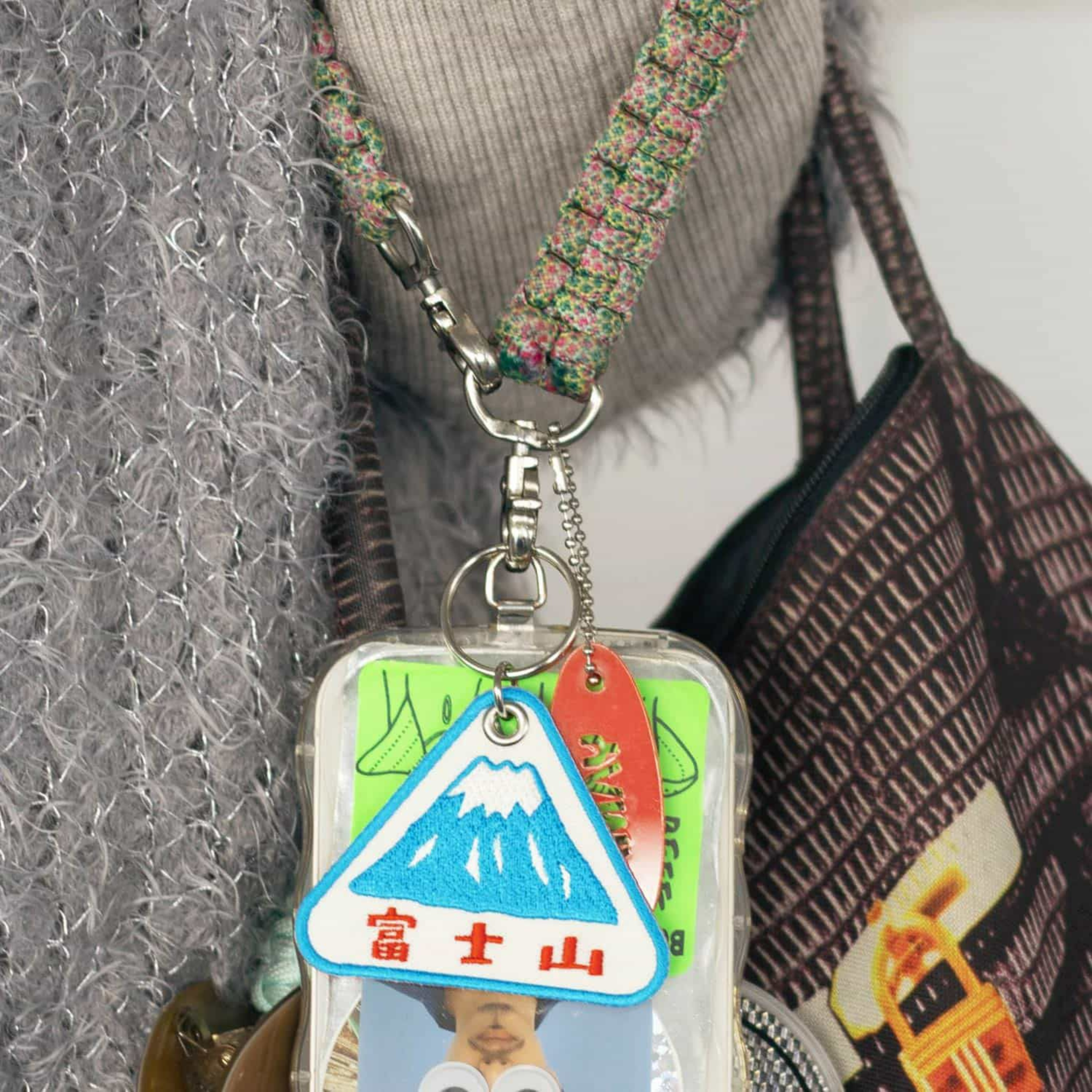 Een close-up van een lanyard met een doorzichtige badgehouder met pasjes en een foto-ID, naast een accessoiretasje dat wordt geaccentueerd door de Hightide Stitch Work Sleutelhanger Fuji van Hightide; een grijze sjaal en tas met patroon op de achtergrond.