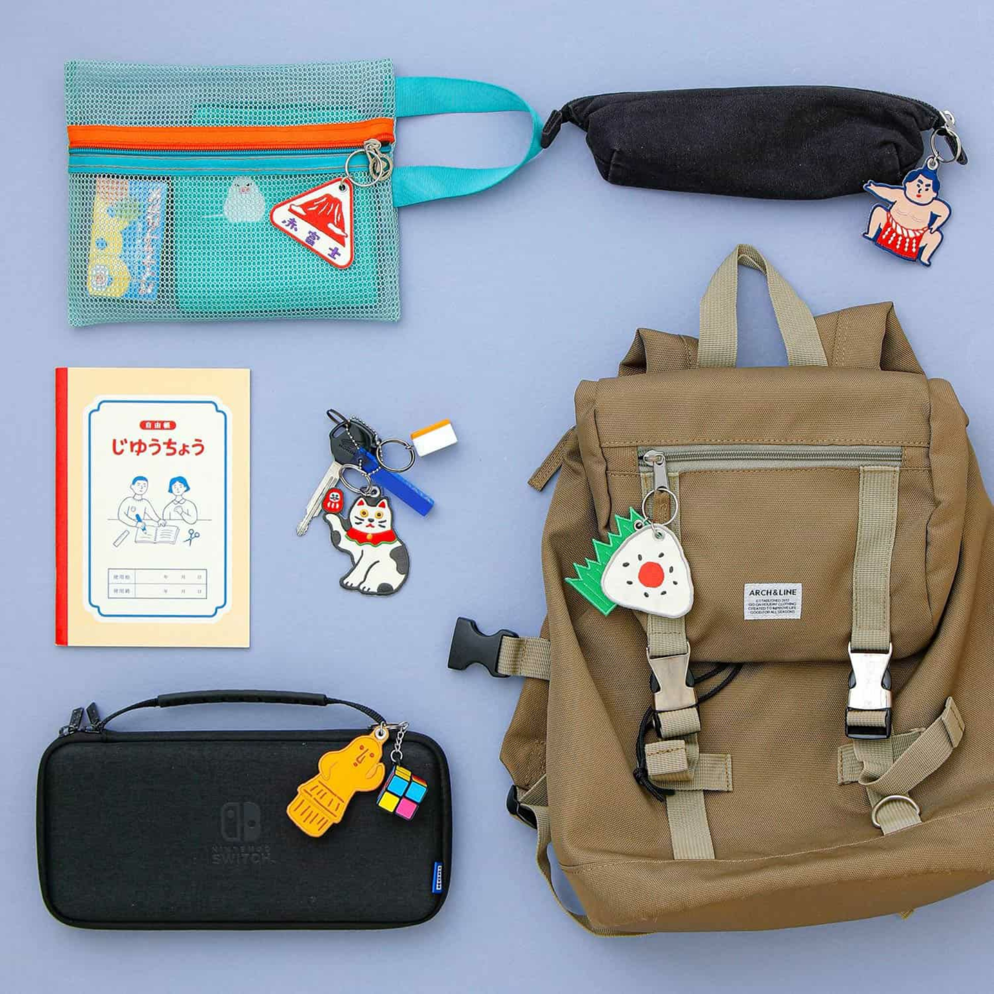 Een platte lay van een bruine rugzak, blauw etui, zwart etui, notitieblok, accessoire tas, Nintendo Switch etui en kleurrijke karakter sleutelhangers zoals de Hightide Stitch Work Sleutelhanger Fuji van Hightide op een blauwe achtergrond.