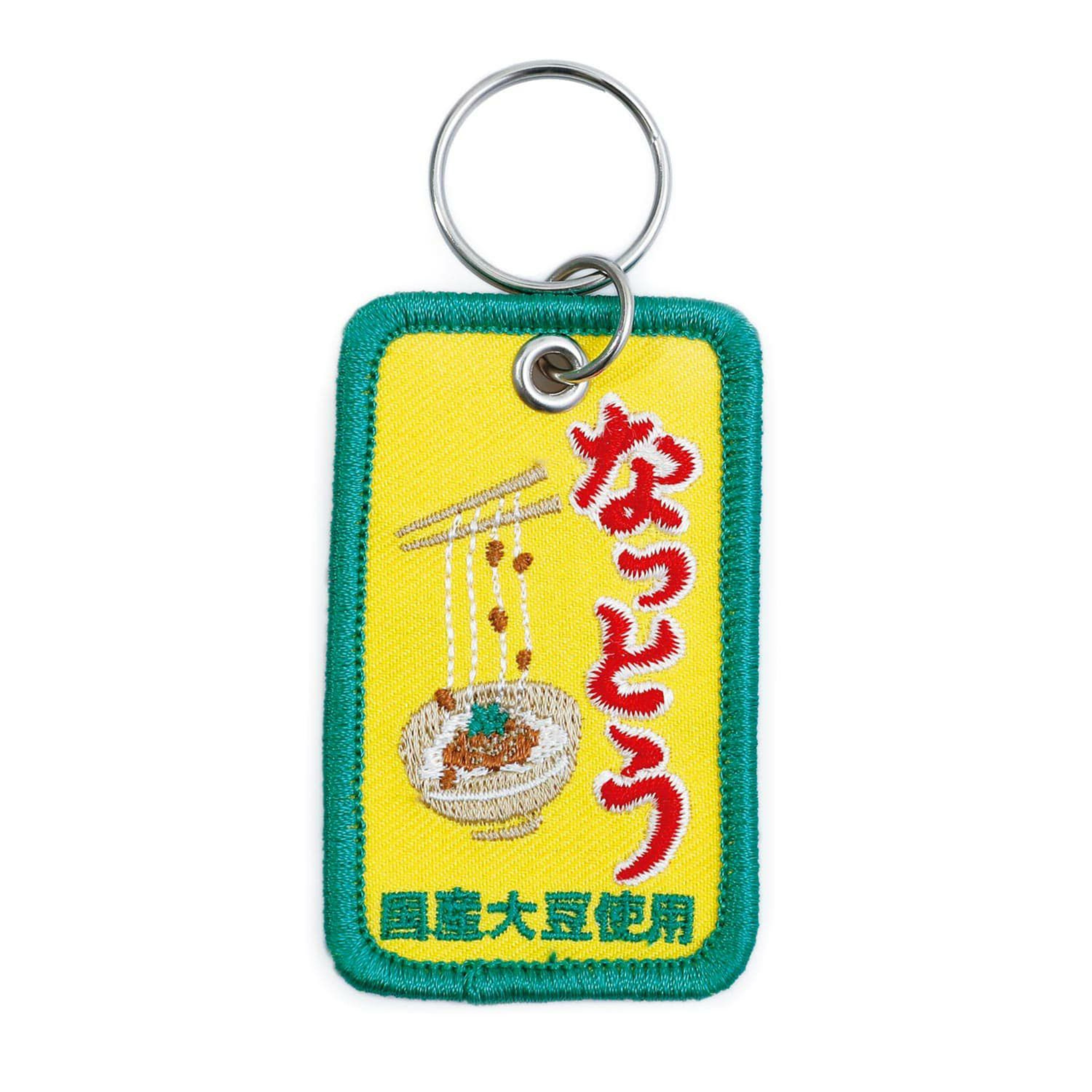 De Hightide Stitch Work Sleutelhanger Natto is een rechthoekige sleutelhanger met een groene rand, met natto en eetstokjes op een gele achtergrond, geaccentueerd door vetgedrukte rode en gele Japanse tekst. Merk: Hightide.