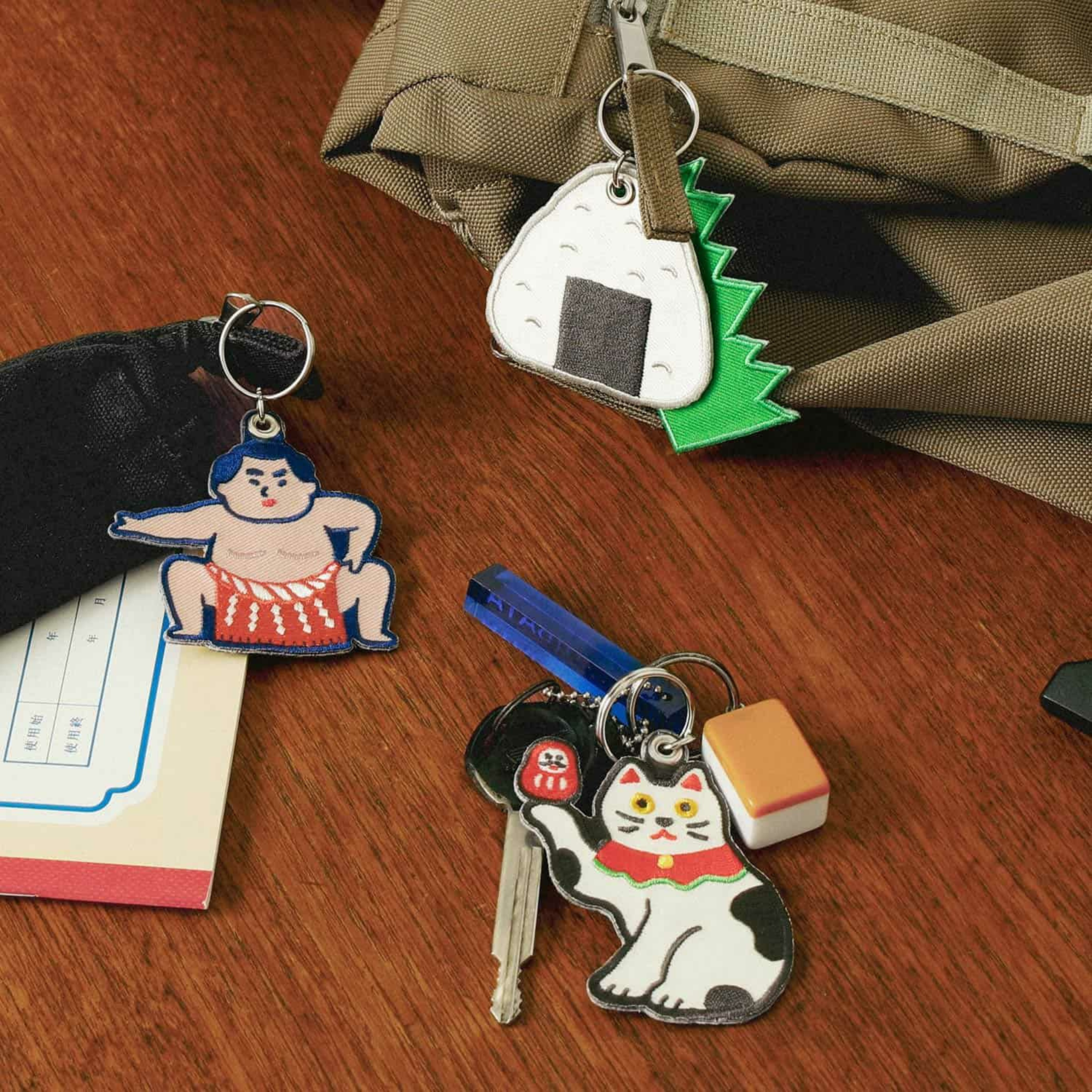 Drie Hightide Stitch Work Sleutelhanger Sumo Worstelaar sleutelhangers van Hightide - een sumoworstelaar, een rijstbal met zeewier en een zwaaiende kat - zijn bevestigd aan sleutels en een tas op een houten oppervlak met kleine voorwerpen in de buurt.