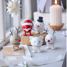 Vier kleine Hoptimist Engel Bumble S Wit beeldjes, geïnspireerd op het Deense ontwerp van Gustav Ehrenreich, staan op een besneeuwd oppervlak naast de Kerstman en een sneeuwpop verlicht door kaarslicht en sterversieringen bij het raam.