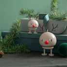 Twee crèmekleurige Hoptimist Rendier Bumble M Latte beeldjes met rode neuzen en gewei staan tussen dennentakken en groene geschenkdozen, ideaal als designobject of Kerstmis decoratie tegen een zachtgroene achtergrond.