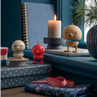 Drie Hoptimist Bumble S Rood beeldjes met verende lichamen staan op decoratieve doosjes naast een kaars, lint en een dennentak in pot, en brengen speels Deens design van Hoptimist naar een blauwe vensterbank.