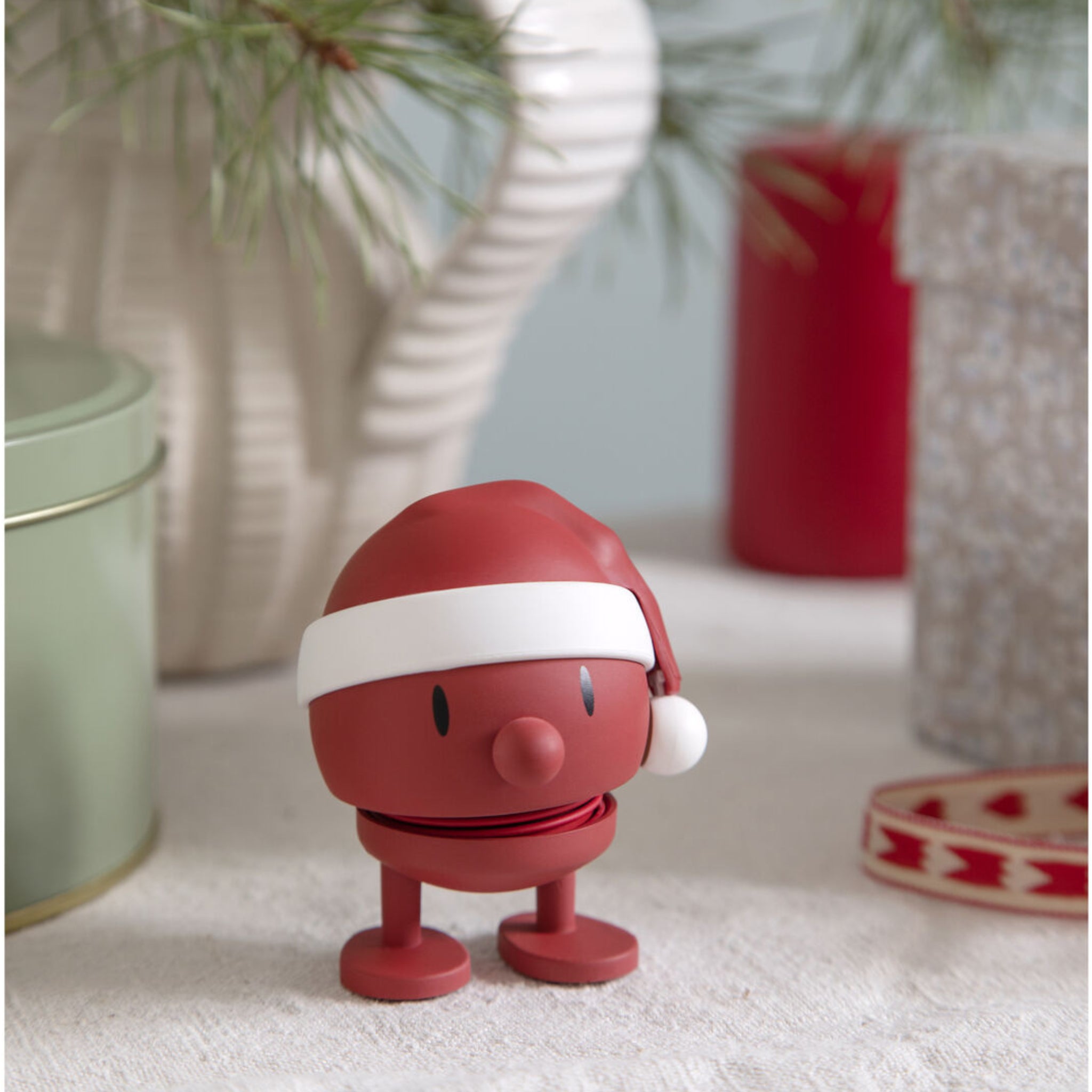 De Hoptimist Soft Santa Bumble S Rood van Hoptimist, een rond rood figuurtje met een kerstmuts, staat op een feestelijke tafel met dennenbomen, cadeautjes en linten - een symbool van Deens design en vakantievreugde.