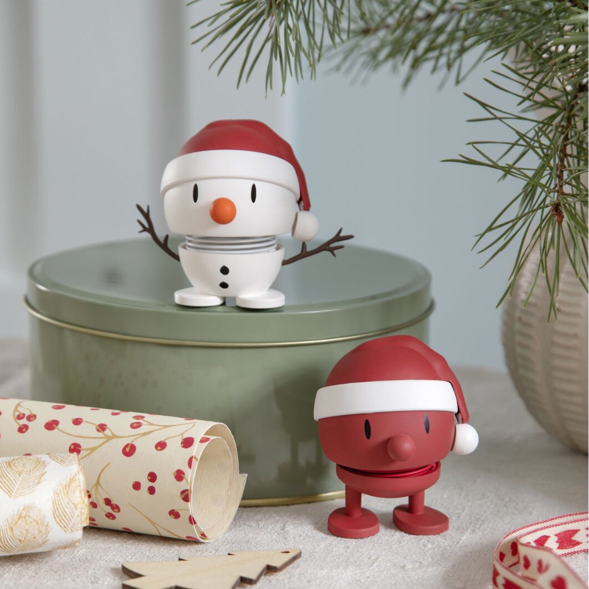 Twee kleine, ronde kerstfiguurtjes - een Hoptimist Soft Santa Bumble S Rood en een vrolijke sneeuwpop - dragen kerstmutsen op een groen blikken doosje bij een dennentak. Ontworpen door Hoptimist, brengen ze een Deens geïnspireerde feestelijke charme in je vakantiedecor.