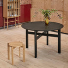 Een klein houten krukje staat naast de Hubsch Fjord Eettafel Essenhout Zwart D120, met daarop een vaas met groen, op een lichte houten vloer in een moderne kamer met houten wanden en een minimalistische inrichting.