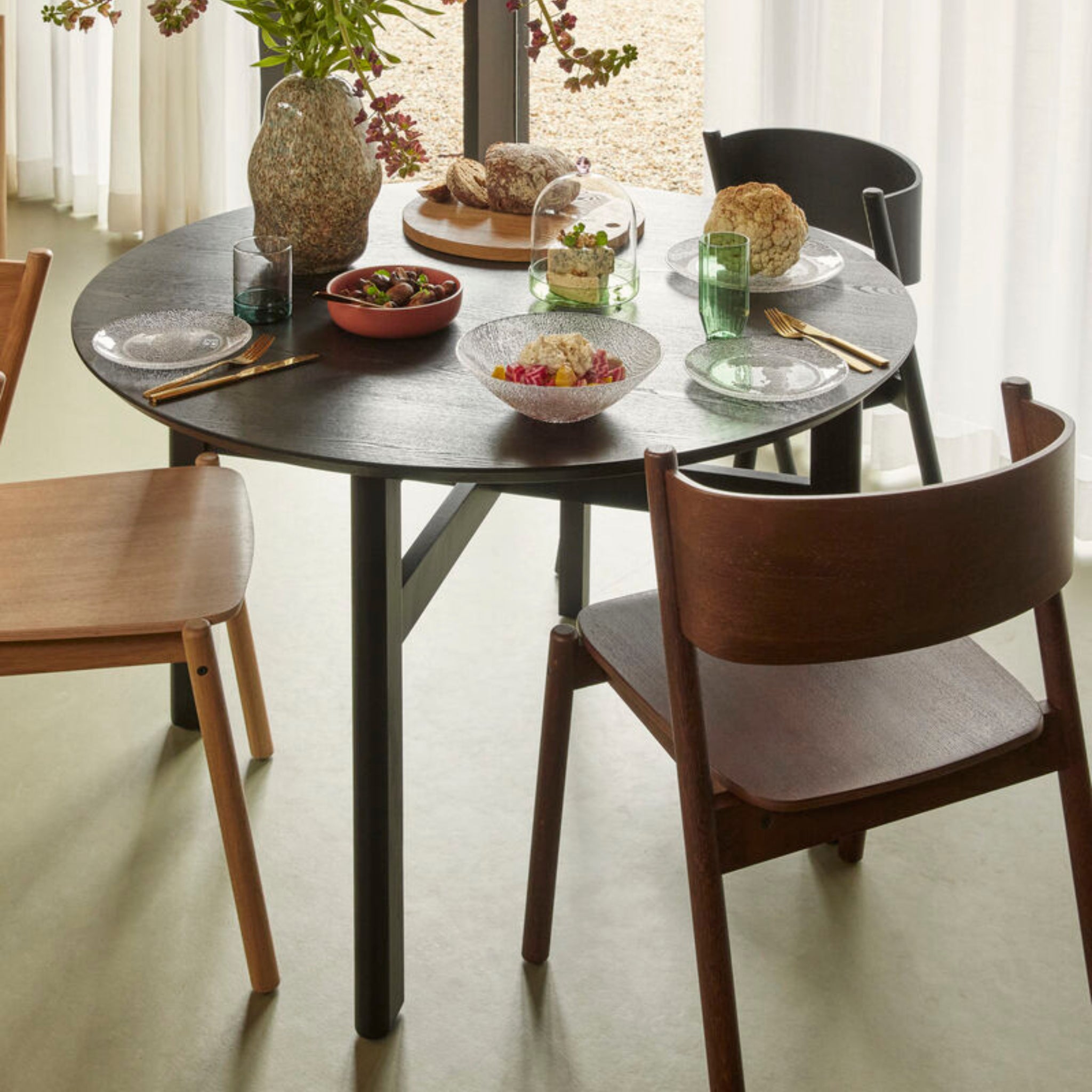 De Hubsch Fjord Eettafel Essenhout Zwart D120 is sfeervol gedekt voor drie personen met borden, glazen, goud bestek en schalen eten. Vazen met bloemen en brood onder een cloche sieren de tafel bij het raam met lichte gordijnen.
