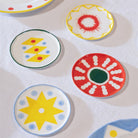 Vier kleine &Klevering Festivo Borden D13 borden (set van 4) van &Klevering, met gedurfde handgeschilderde gele sterren, rode zonnestralen en speelse stippen, worden weergegeven op een wit tafelkleed voor een levendige look.