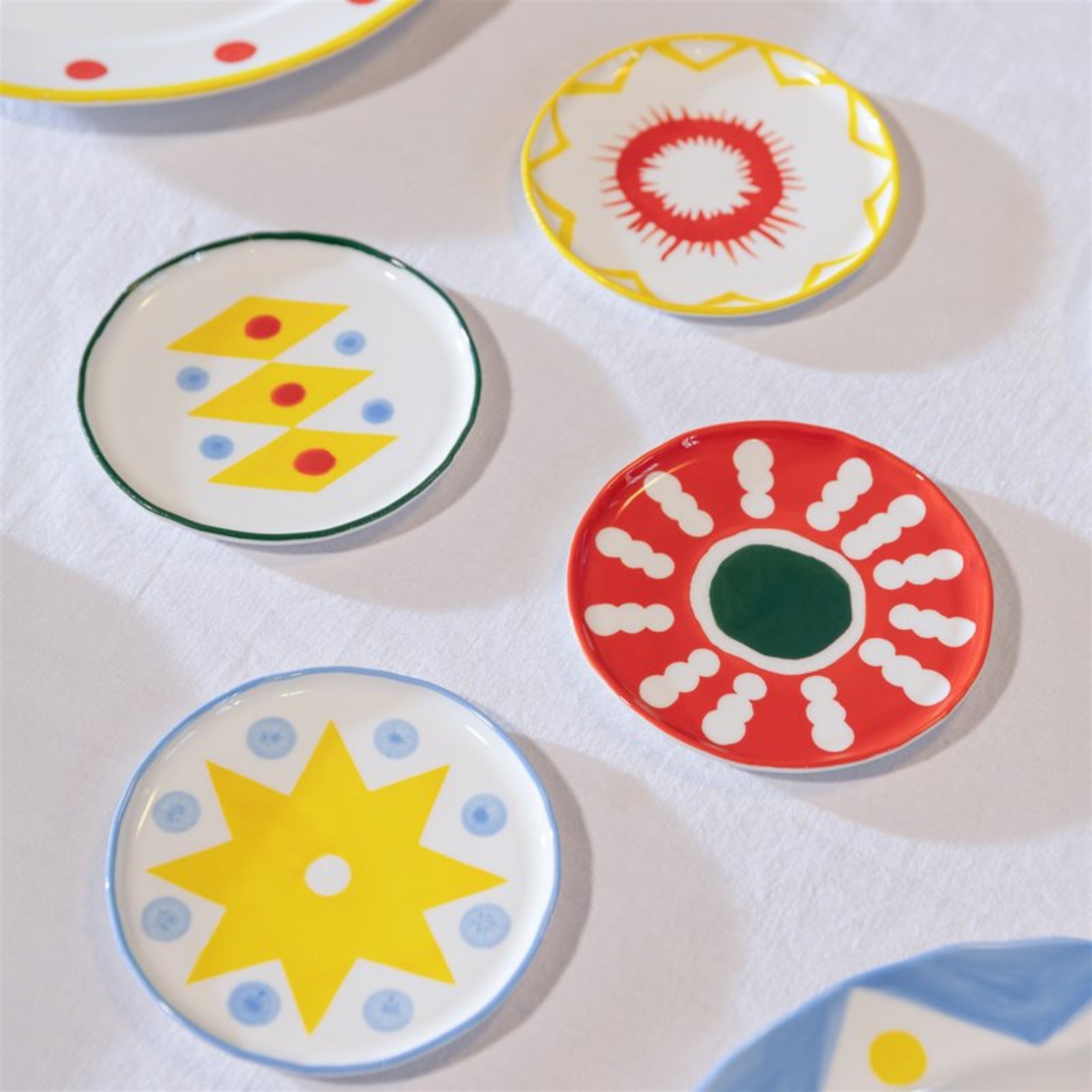 Vier kleine &Klevering Festivo Borden D13 borden (set van 4) van &Klevering, met gedurfde handgeschilderde gele sterren, rode zonnestralen en speelse stippen, worden weergegeven op een wit tafelkleed voor een levendige look.