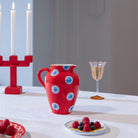 Een &Klevering Festivo Kan Rood 1,5L met blauwe en witte cirkels rust op een wit tafelkleed naast een bord bessen, een champagneglas en een rode kandelaar met drie brandende kaarsen, die samen een charmante decoratieve display vormen.