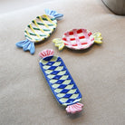 Drie &Klevering Goody Bord 27/11 keramische schalen van &Klevering, in de vorm van ingepakte snoepjes met rode, blauwe, gele en groene ruitjespatronen, staan op beige stof - perfect als speels middelpunt.