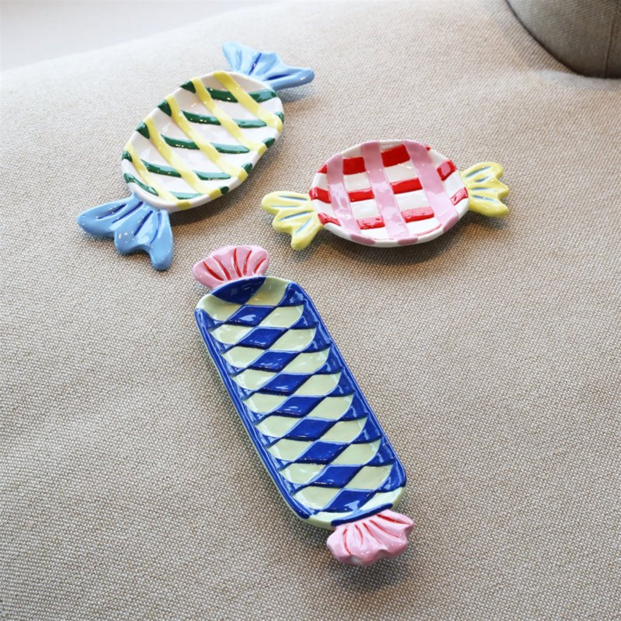 Drie &Klevering Goody Bord 27/11 keramische schalen van &Klevering, in de vorm van ingepakte snoepjes met rode, blauwe, gele en groene ruitjespatronen, staan op beige stof - perfect als speels middelpunt.