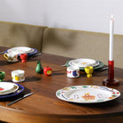 Een houten eettafel gedekt met kleurrijke borden, waaronder de &Klevering Haines Schotel 35/26. Kleine fruitige decoraties en een hoge kaars in een rode houder voegen charme toe, terwijl een beige bank met kussens op de achtergrond staat.