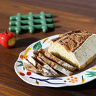 Op de &Klevering Haines Schotel 35/26 van &Klevering ligt een gedeeltelijk aangesneden rustiek brood met bloemmotieven. Vlakbij liggen een kleine rode appel en een groen onderzettertje op de houten tafel.