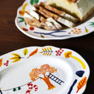 Een close-up van twee &Klevering Haines Schotel 28/22 schotels van &Klevering, een met gesneden brood en een leeg, beide versierd met kleurrijke, grillige illustraties - een ideaal uniek cadeau op een donkere houten tafel.