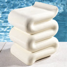 De &Klevering Whip Pillar Bijzettafeltje Gebroken Wit heeft een golvend, gestapeld ontwerp en een crèmekleur. Het combineert sculpturale flair met duurzame glasvezel - perfect naast een zwembad op een licht stenen oppervlak.