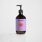 De bruine pompflacon van Matriks Fragrances Hand & Body Wash Amber & Patchouli 490 ml staat op een wit oppervlak. Het lichtpaarse etiket met cirkelvormig design benadrukt deze veganistische, stijlvolle en geurige body wash.
