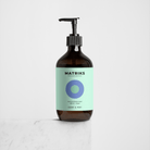 Een bruine pompflacon van Matriks Fragrances Hand & Body Wash Cedar & Mint 490 ml met een lichtblauw etiket staat op wit marmer, met een veganistische formule ideaal als handzeep of body wash tegen een effen grijze achtergrond.