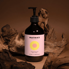 Een 490 ml fles Matriks Fragrances Hand & Body Wash Citrus & Grapefruit, met een roze etiket, rust tussen drijfhout op een bruine ondergrond, wat de frisse geur van grapefruit oproept in deze veganistische wash.