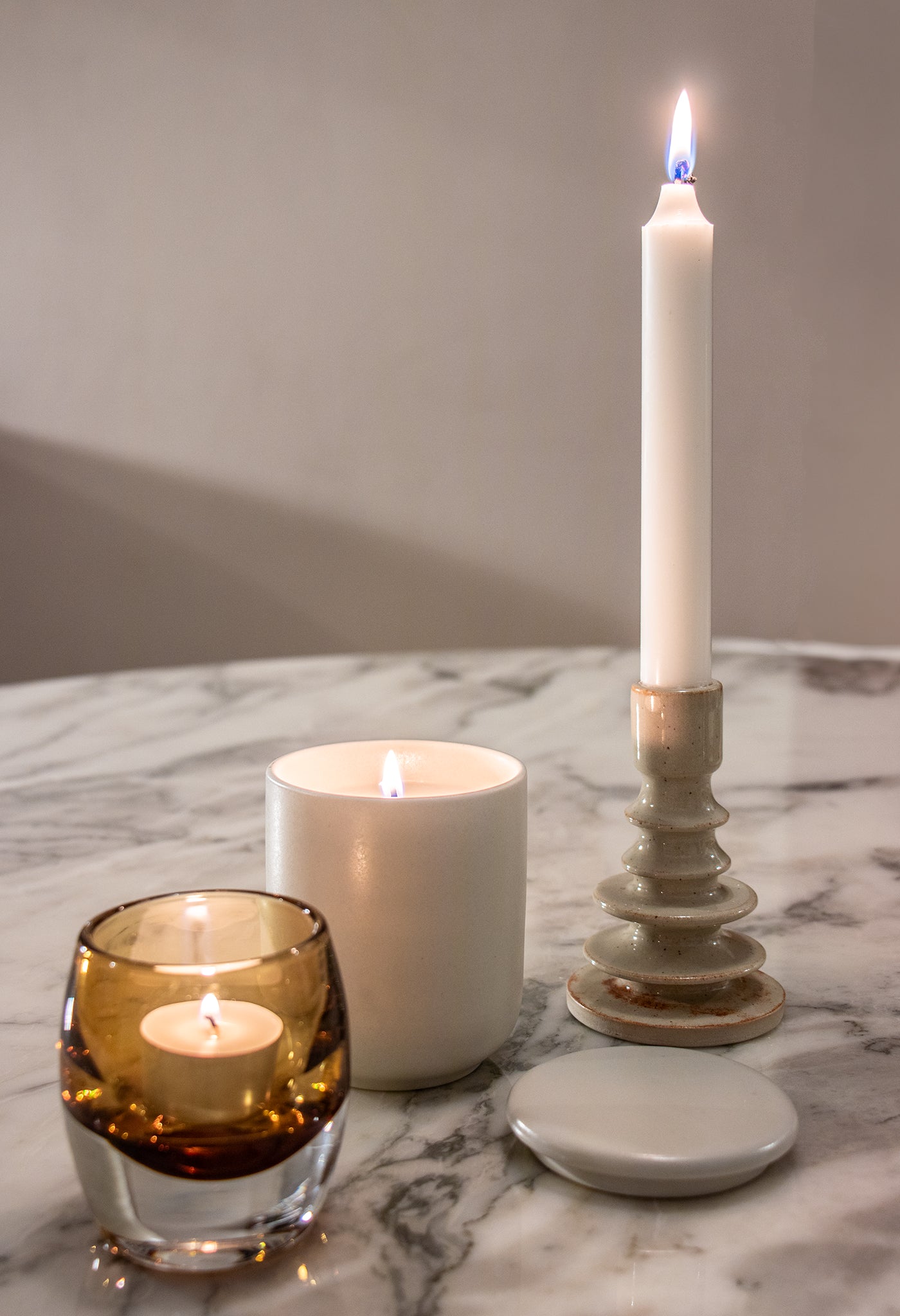 Drie brandende geurkaarsen van Matriks Fragrances zitten op marmer: de handgemaakte Keramiek Woodland Pine 270g kaars, een witte kaars in een wit potje en een waxinelichtje in bruin glas. Allemaal gemaakt in Antwerpen. Een wit deksel rust vlakbij.