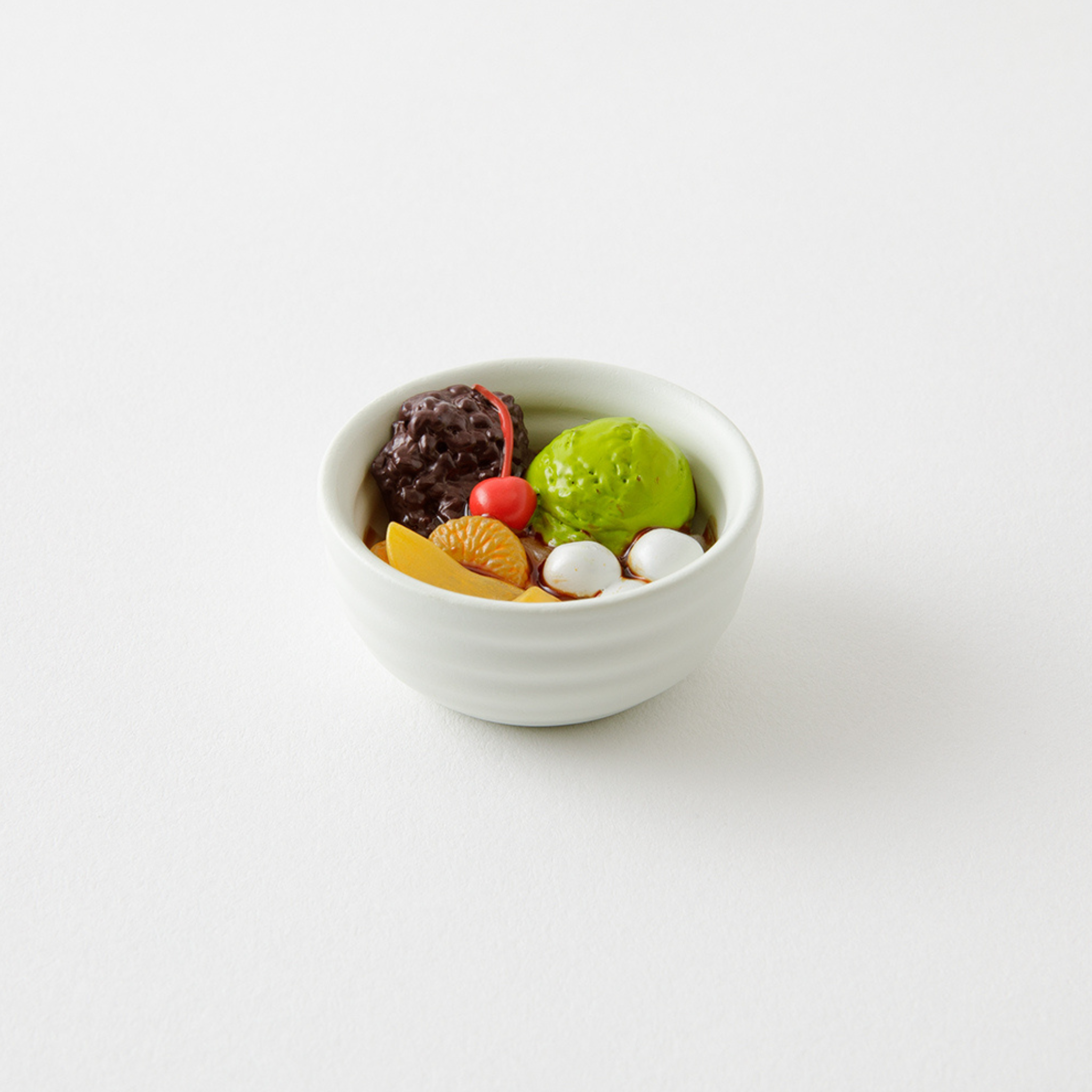 Een kleine witte schaal gevuld met kleurrijk dessert in Japanse stijl - groen matcha-ijs, plakjes fruit, rode bonenpasta, mochibolletjes en een kers - maakt van de Midori Miniatuur Magneet Anmitsu van Midori een verrukkelijke verzamelmagneet.
