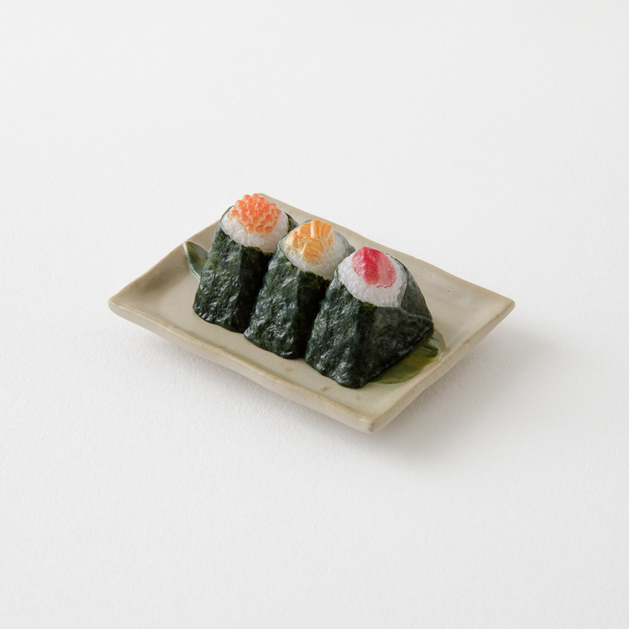 Drie stukjes sushi, verpakt in zeewier en belegd met kuit, zalm en tonijn, staan prachtig uitgestald op een keramisch bord. Dit is de Midori Miniatuur Magneet Onigiri voor je koelkast!.