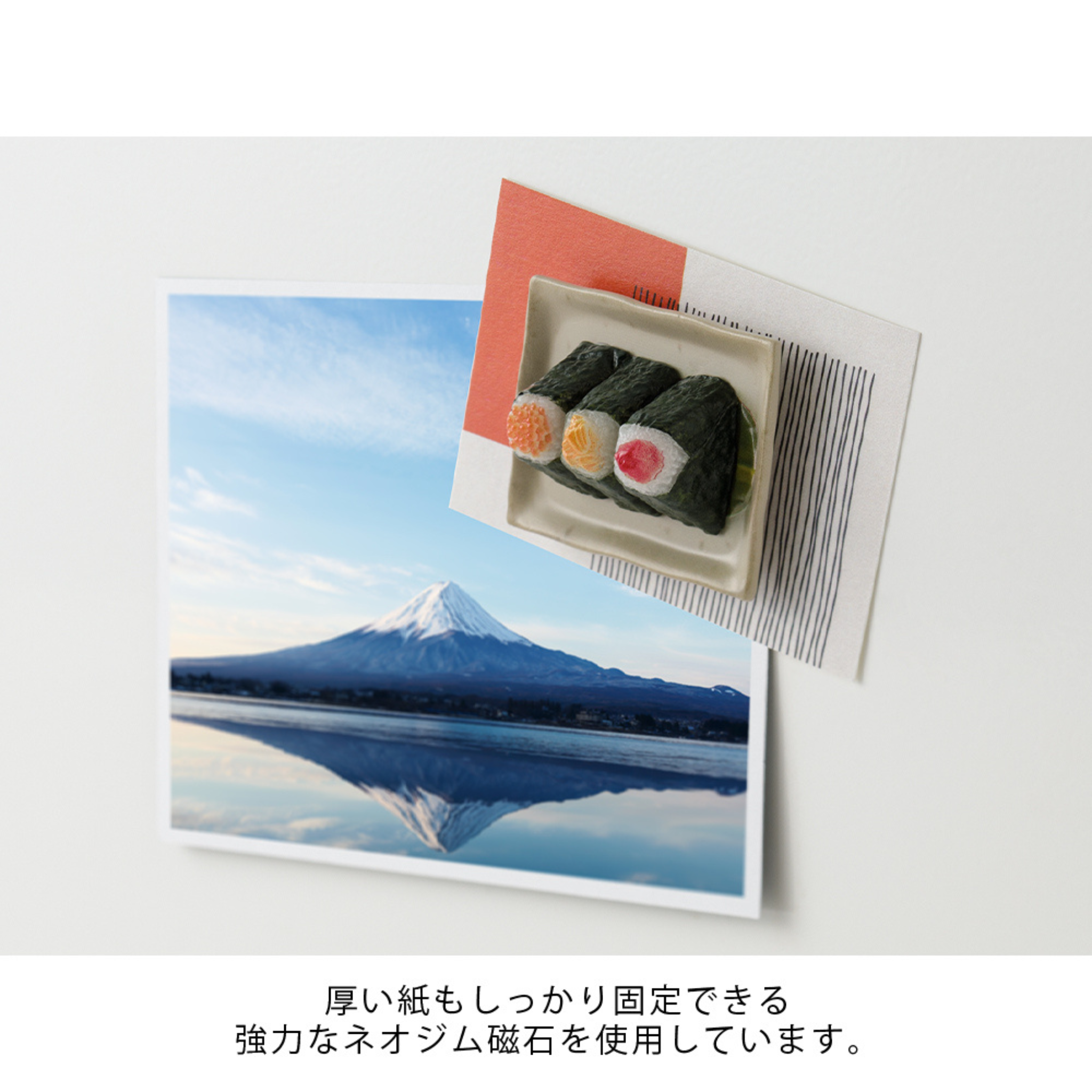 Een foto van Mt. Fuji weerspiegeld in een meer is vastgepind op een wit oppervlak met de Midori Miniatuur Magneet Onigiri, een leuke sushi-vormige koelkast magneet van Midori, die een briefje vasthoudt. Onder de foto staat Japanse tekst.