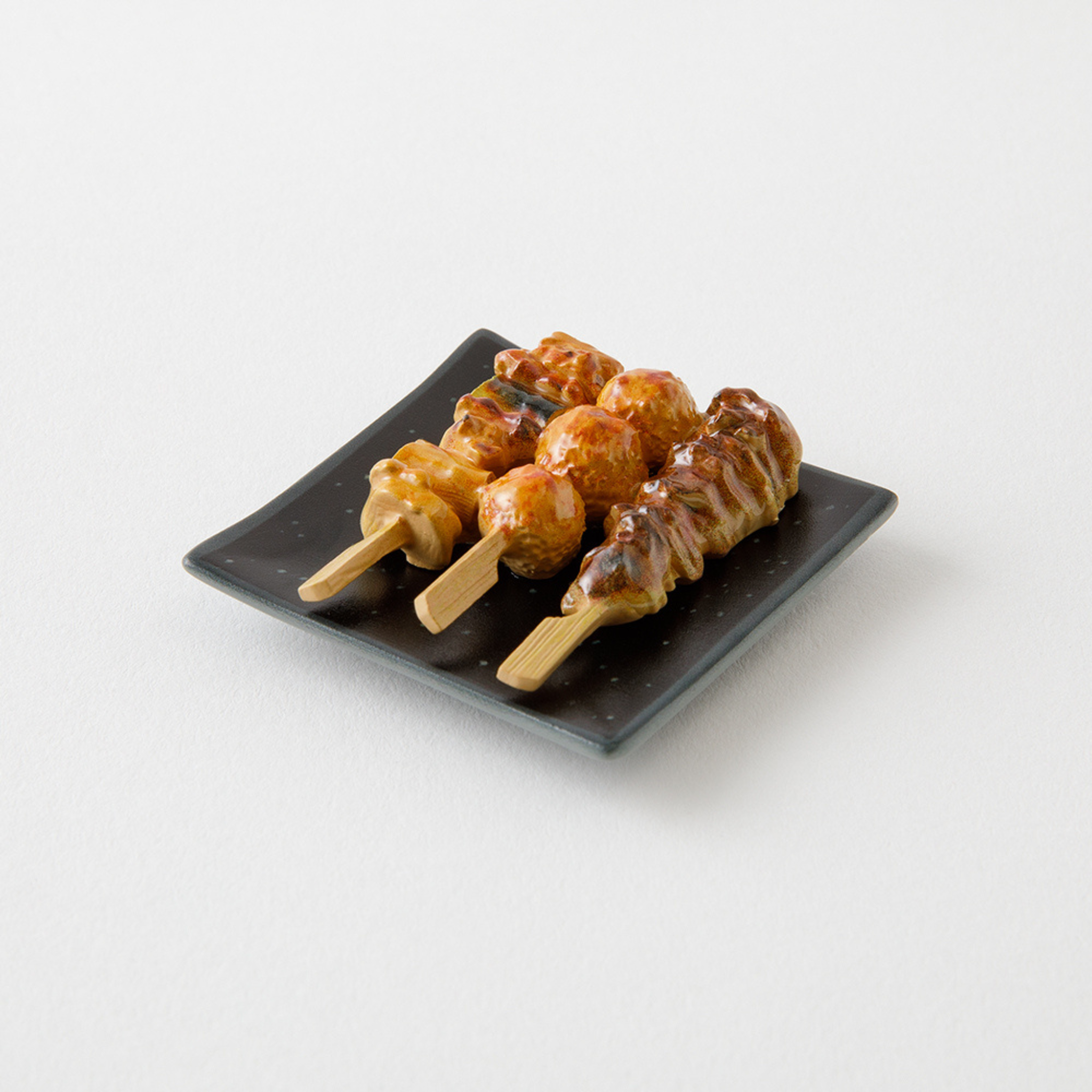 De Midori Miniatuur Magneet Yakitori bestaat uit drie gegrilde kipspiesjes met saus op een zwart bord - ideaal om je koelkast mee op te sieren en perfect voor fans van Japanse keuken.