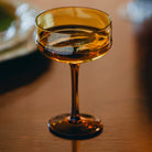 Een close-up van de Paveau Swirl Champagne Coupe Dune Geel 26 cl van Paveau, gemaakt van borosilicaatglas, staat leeg op een houten tafel met zacht licht dat weerkaatst op het amberkleurige oppervlak.