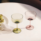 Twee lege Paveau Swirl Champagne Coupe Silt Bordeaux glazen (26 cl) van Paveau - een groene, een roze - staan op een wit tafelkleed naast een bord met een groene bloem en een gevouwen servet, verlicht door zacht natuurlijk licht.