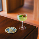 Een groene cocktail gegarneerd met gedroogde limoen en basilicum staat op een houten tafel naast een geweven onderzetter. Geserveerd in de Paveau Swirl Champagne Coupe Wave Helder 26 cl van Paveau, wordt de frisheid versterkt door de warme, wazige achtergrond.
