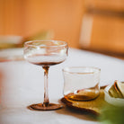 Een Paveau Swirl Champagne Coupe Glow Beige 26 cl siert een wit tafelkleed, vergezeld door een korte tumbler, geweven placemat en gevouwen servet. De zacht vervaagde achtergrond baadt het tafereel in een warme gloed van Paveau.