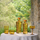 Paveau amberkleurig glaswerk, waaronder de Paveau Swirl Waterglas Dune Geel 24 cl, flessen, tumblers en een coupeglas, staat op een ronde tafel met een wit kleed. Weelderig groen gebladerte en een stenen muur verschijnen op de achtergrond.