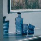 Een Paveau Swirl Waterglas Midnight Blauw 24 cl van Paveau staat op een blauwe richel, het middernachtblauwe glas werpt golvende schaduwen. Daarachter siert een schilderij met diepgroene, gestructureerde penseelstreken de muur.