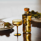 Een Paveau Swirl Champagne Coupe Dune Geel 26 cl van Paveau staat op een gepolijste tafel, in combinatie met een bijpassend glas. Borden met eten en groene decoraties creëren een zacht vage achtergrond.