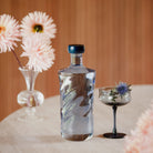 De Paveau Swirl Waterfles Midnight Blauw 1,25 l van Paveau staat op een ronde tafel naast een zwartgesteeld cocktailglas met blauwe distel en een vaas bleekroze bloemen, met een houten achtergrond.