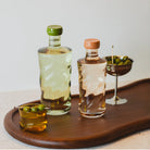 Twee borosilicaatglazen flessen met groene en perzikkleurige doppen bevatten heldere en amberkleurige vloeistoffen op een houten dienblad. Een metalen beker en de Paveau Swirl Waterglas Dune Geel 24 cl van Paveau, beide met groene olijven, maken het elegante tafereel compleet.