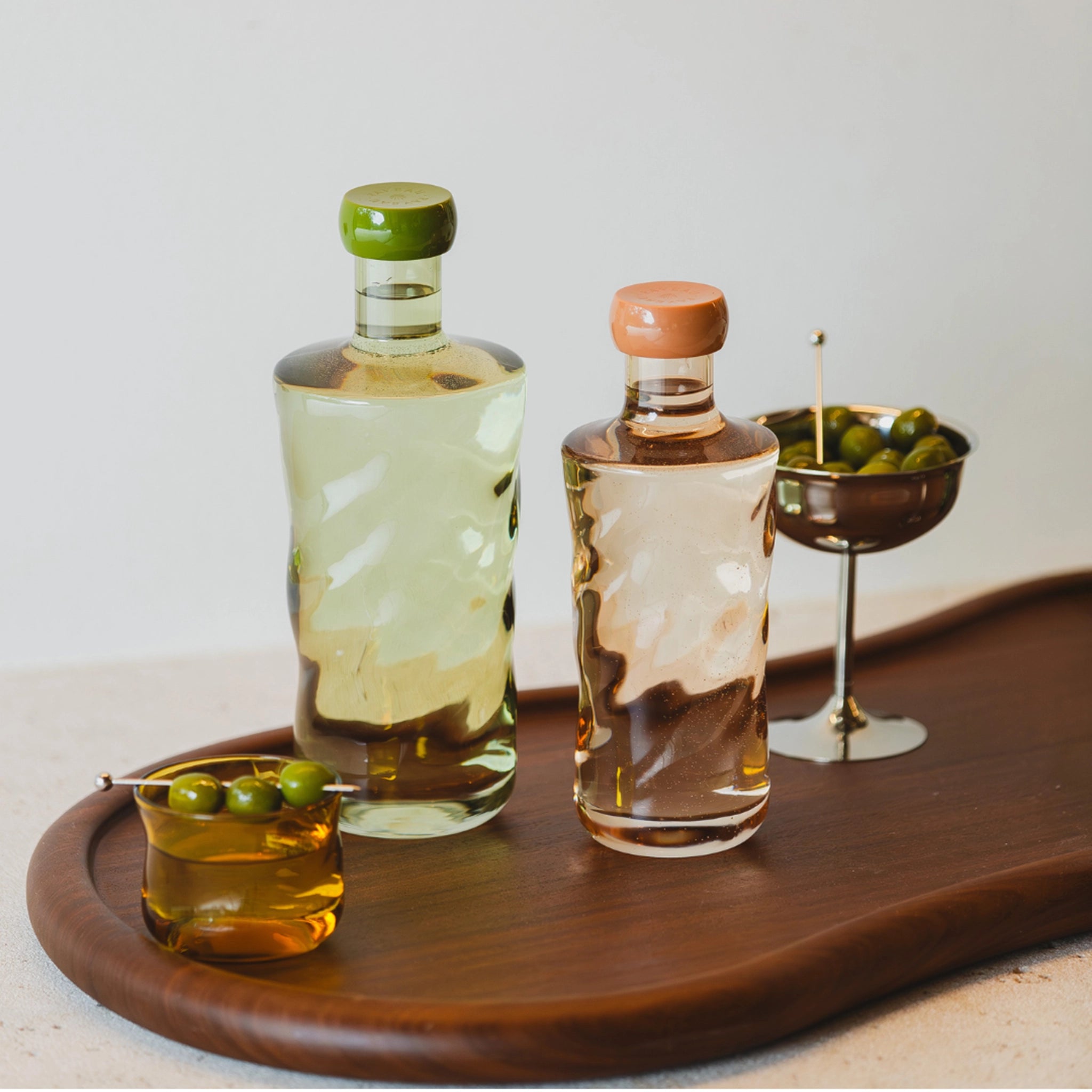 Twee borosilicaatglazen flessen met groene en perzikkleurige doppen bevatten heldere en amberkleurige vloeistoffen op een houten dienblad. Een metalen beker en de Paveau Swirl Waterglas Dune Geel 24 cl van Paveau, beide met groene olijven, maken het elegante tafereel compleet.