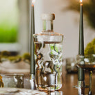 De Paveau Swirl Waterfles Wave Helder 0,75 l van Paveau met witte dop en textuur staat op een houten eettafel naast twee brandende kaarsen, met een vage groene achtergrond.