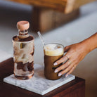 Een hand met ringen reikt naar ijskoffie met een rietje naast een heldere fles - beide op marmer bovenop hout. De Paveau Swirl Longdrink Glas Glow Beige 46 cl van Paveau voegt elegantie toe met zijn ontwerp van borosilicaatglas.