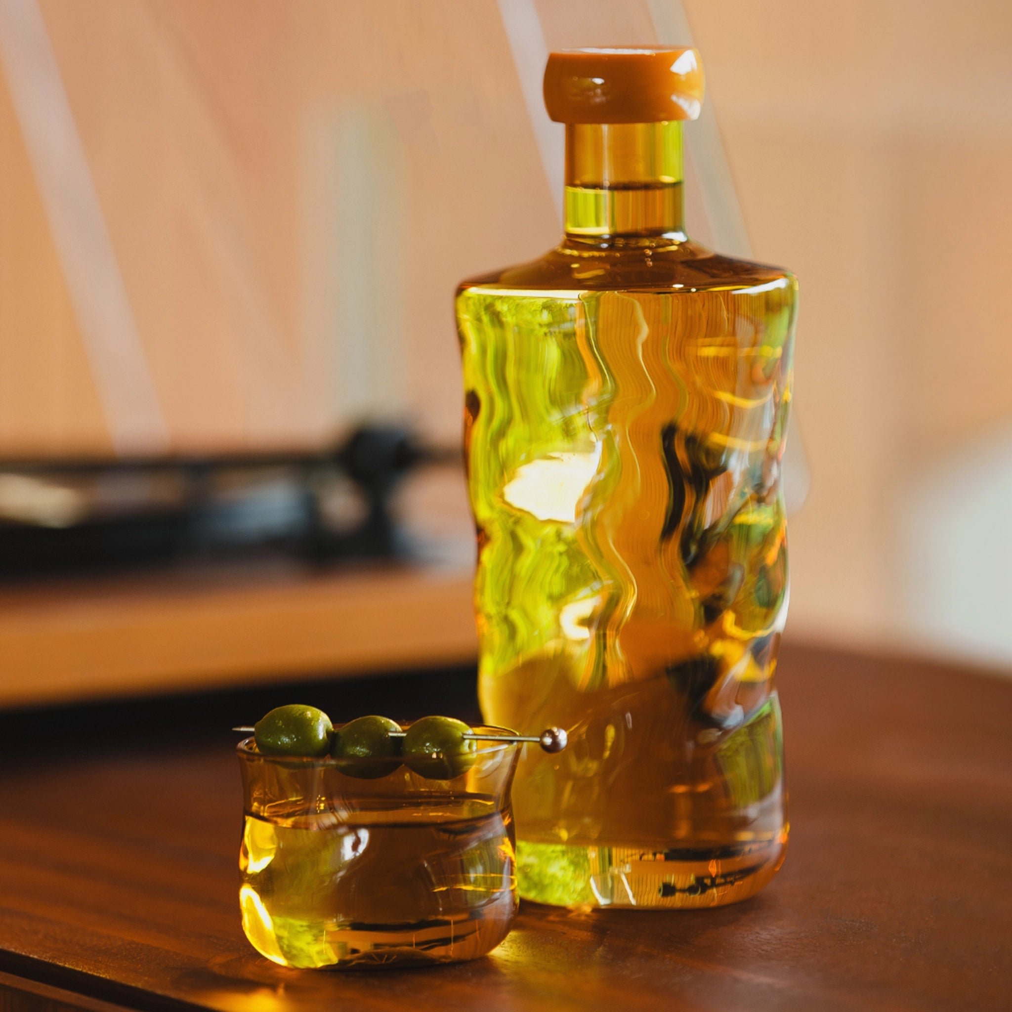 Een Paveau Swirl Waterglas Dune Geel 24 cl van Paveau, gevuld met gouden vloeistof, staat op een houten oppervlak naast een schaaltje groene olijven. De zacht vervaagde achtergrond creëert een elegante, op duinen geïnspireerde sfeer.
